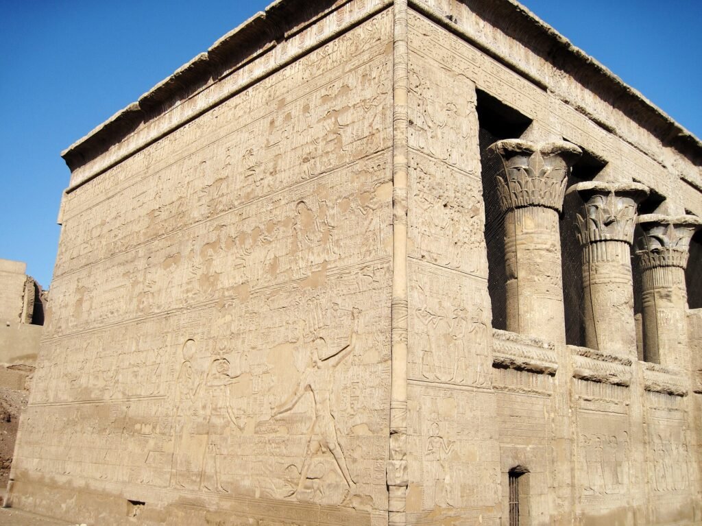 La parete esterna scolpita del tempio di Esna nella luce bassa del pomeriggio, con i capitelli floreali della sala ipostila sullo sfondo