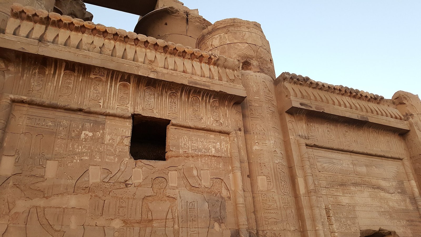 La parete scolpita del tempio di Kom Ombo con la cornice a gola egizia e le colonne sullo sfondo, nella luce calda del tardo pomeriggio