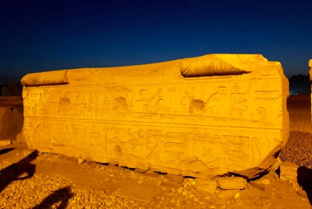 Un grande architrave scolpito caduto a terra fra le rovine di Kom Ombo, illuminato di notte contro il cielo blu