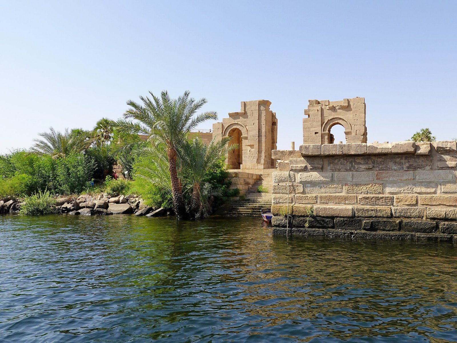 L'isola di Philae vista dall'acqua, con la banchina in pietra, le palme e i resti di un portale ad arco