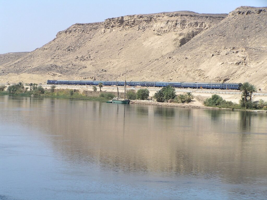 Un treno passeggeri egiziano che costeggia il Nilo fra Assuan e Luxor, con una feluca ormeggiata sulla riva e la parete desertica alle spalle