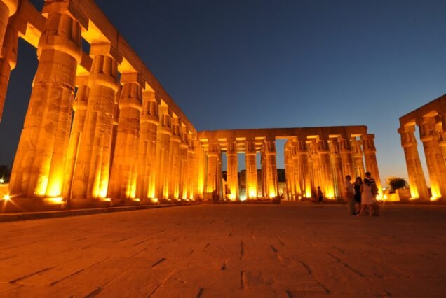 Il colonnato del tempio di Luxor illuminato di notte, con il cielo blu del crepuscolo e alcuni visitatori in lontananza
