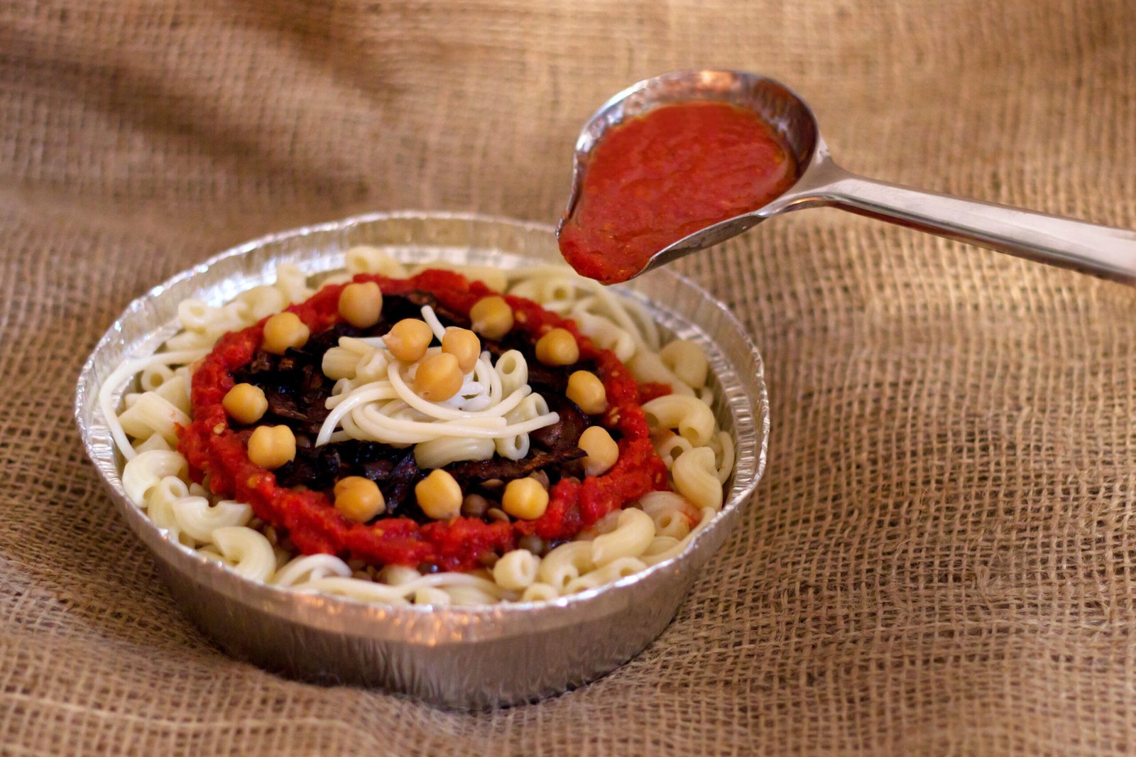 Ciotola di koshari con il mestolo di salsa di pomodoro su tela di iuta