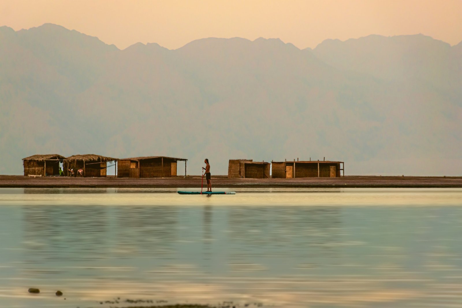 Persona su tavola da paddle al tramonto sulla Blue Lagoon di Dahab davanti a capanne di canne