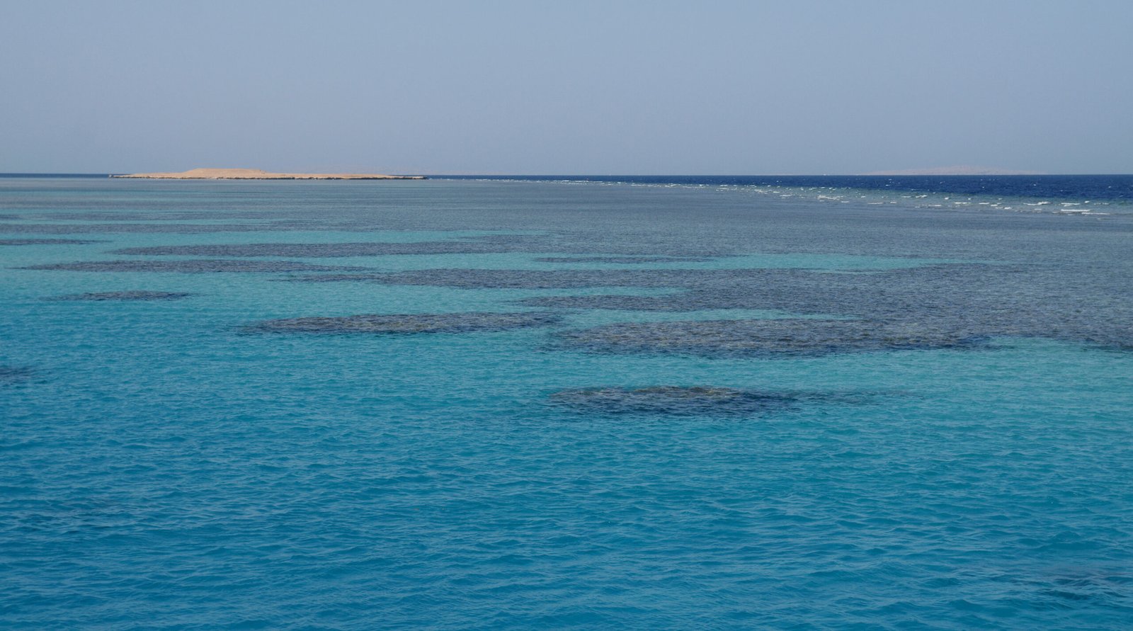 Acque turchesi del Mar Rosso con banchi corallini affioranti e un'isola sabbiosa all'orizzonte