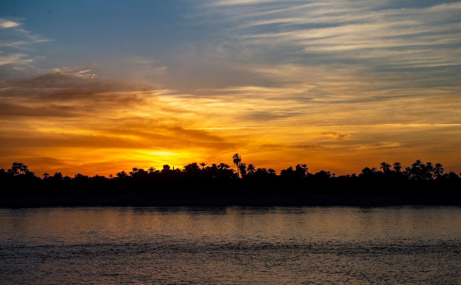 Tramonto dorato sul Nilo con le palme della riva opposta in controluce e il fiume calmo