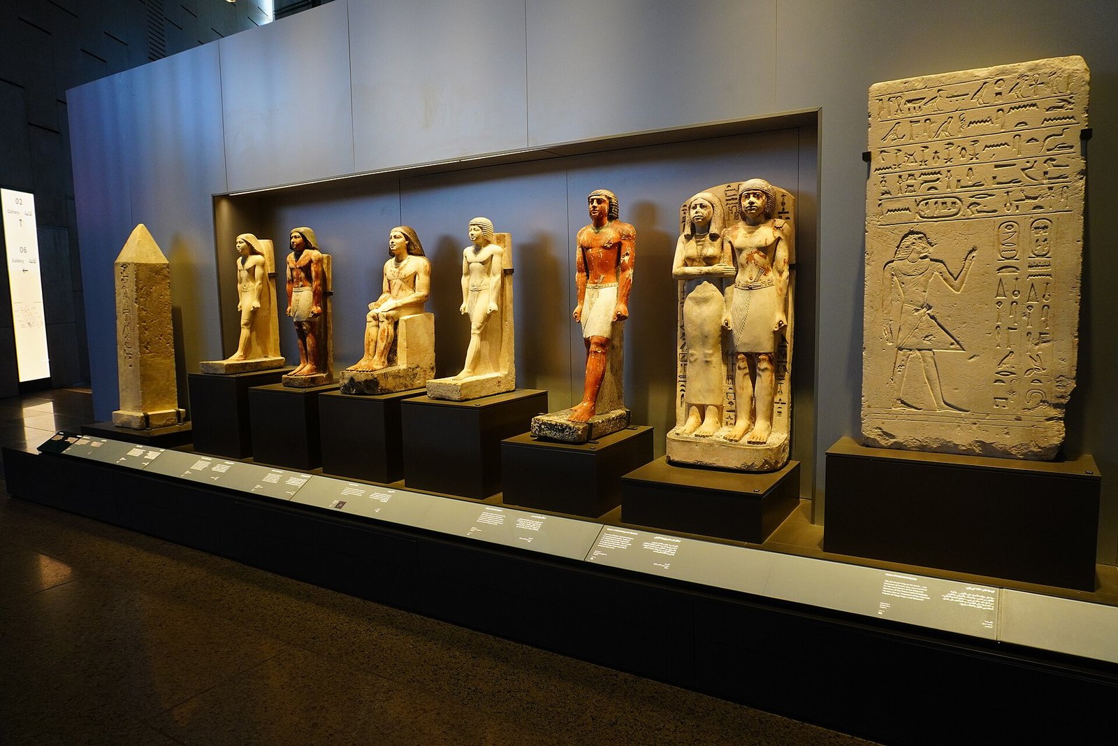 Galleria del Grande Museo Egizio con una fila di statue egizie in pietra su basamenti scuri, un piccolo obelisco sulla sinistra e una stele con geroglifici sulla destra