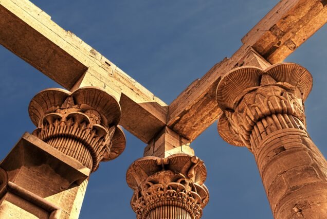 Tre capitelli compositi diversi del tempio di Philae visti dal basso contro il cielo blu, con palma, papiro e loto scolpiti nell'arenaria