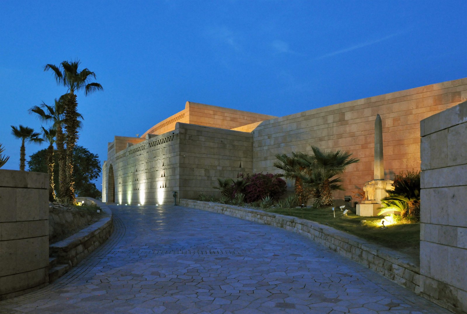 L'ingresso del Museo Nubiano di Assuan al crepuscolo, con il muro di pietra, le palme e un piccolo obelisco illuminati