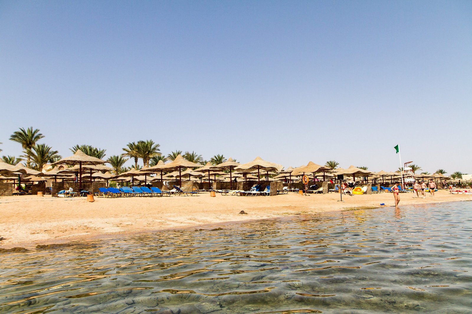 La spiaggia di Hurghada con gli ombrelloni di paglia sotto le palme e l'acqua bassa e limpida