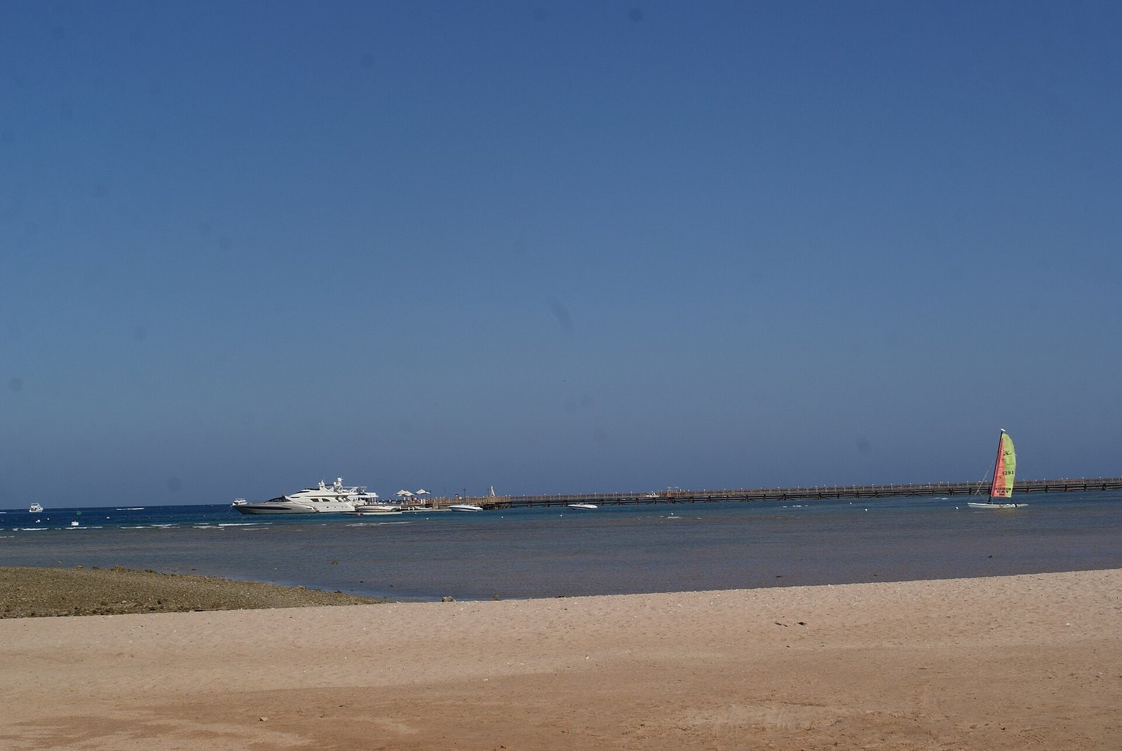 La spiaggia di sabbia di Makadi Bay con il lungo pontile, uno yacht ormeggiato e una barca a vela nel mare calmo