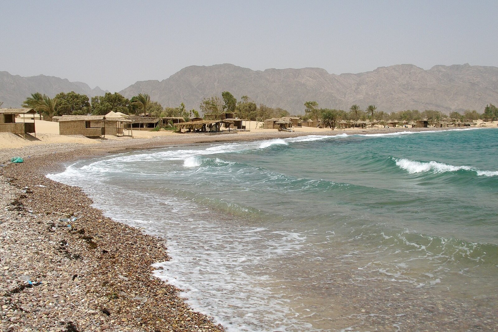 La spiaggia di ciottoli di Nuweiba sul Mar Rosso, con le onde, le capanne di canne e le montagne del Sinai
