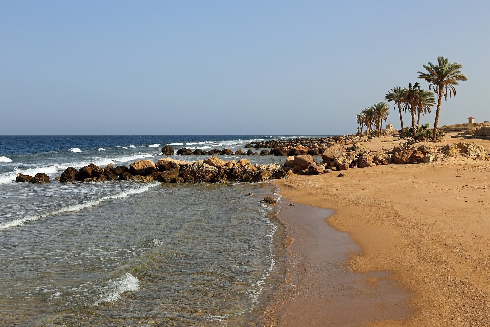 Una piccola baia rocciosa di Sahl Hasheesh con le onde sulla sabbia dorata e le palme sul promontorio